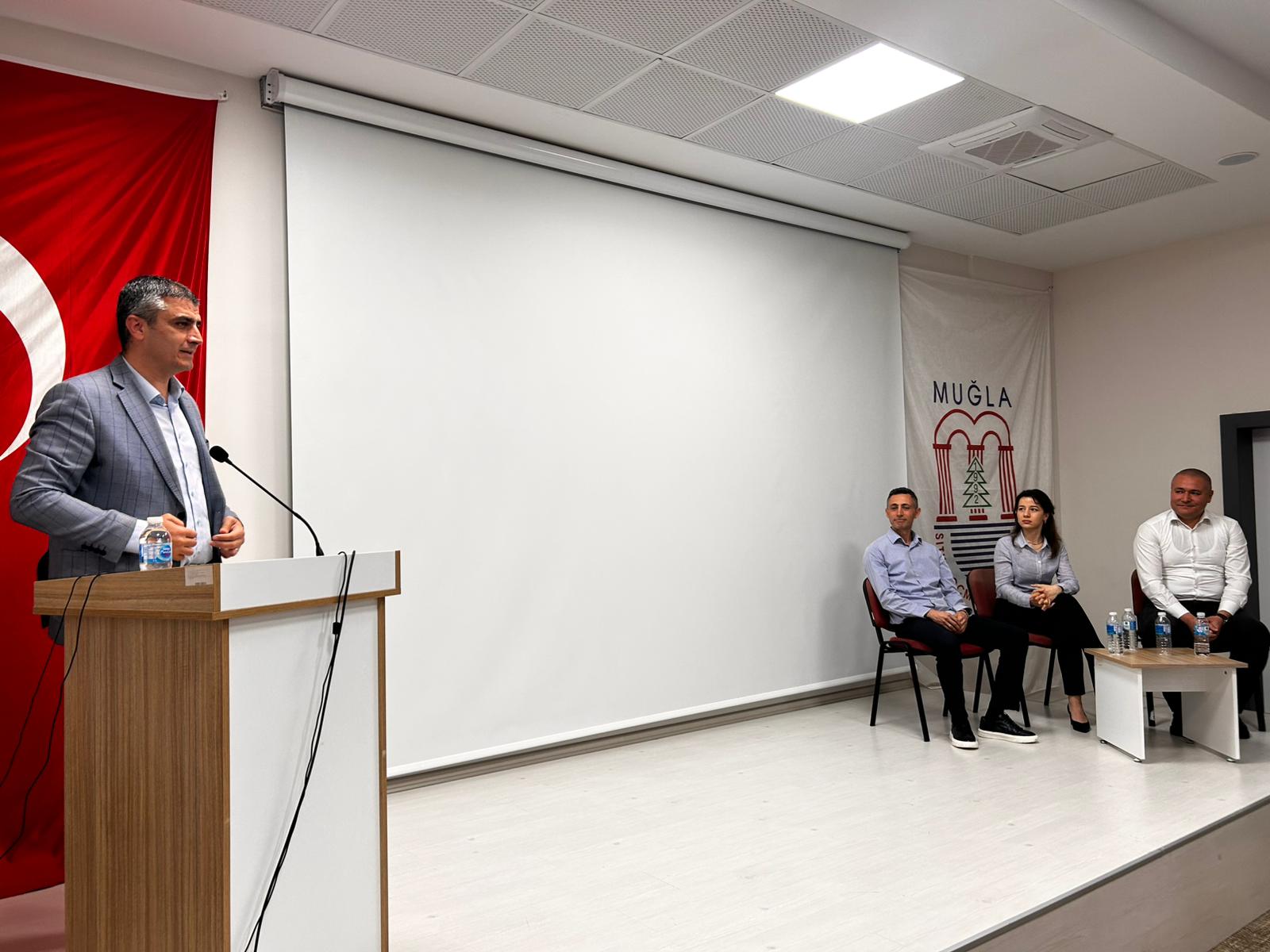 Bir diğer konuşmacının kürsüde olduğu bu karede, panelistlerin sahnenin yan tarafında oturarak sunumu takip ettikleri görülüyor. Muğla Sıtkı Koçman Üniversitesi amblemli flamanın önünde gerçekleşen bu an, etkinliğin akademik aidiyetini ve profesyonel organizasyon yapısını simgeliyor.